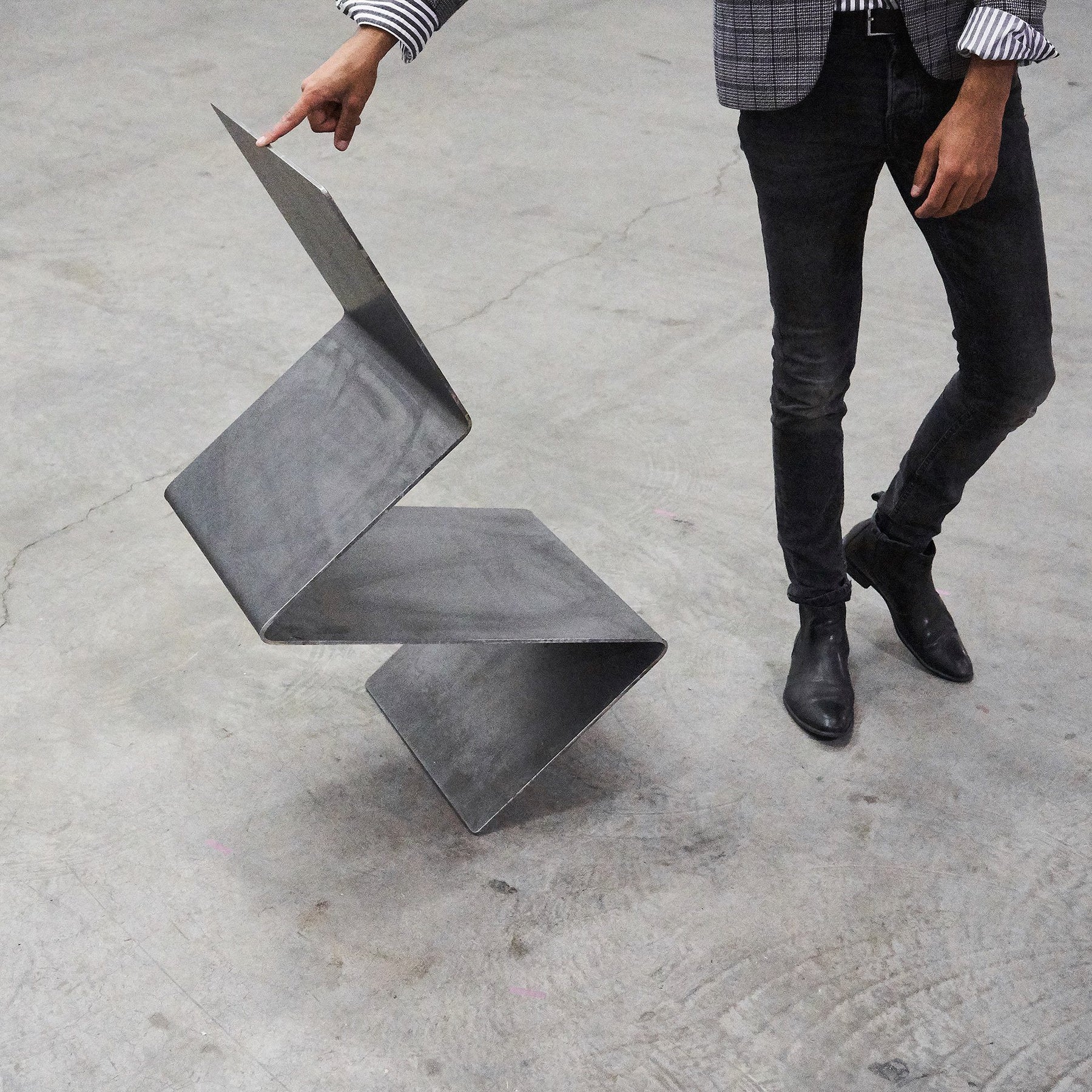 Eine Person kippt mit dem Zeigefinger einen Freischwinger aus rohem Flachstahl, der auf einem Betonboden steht, um