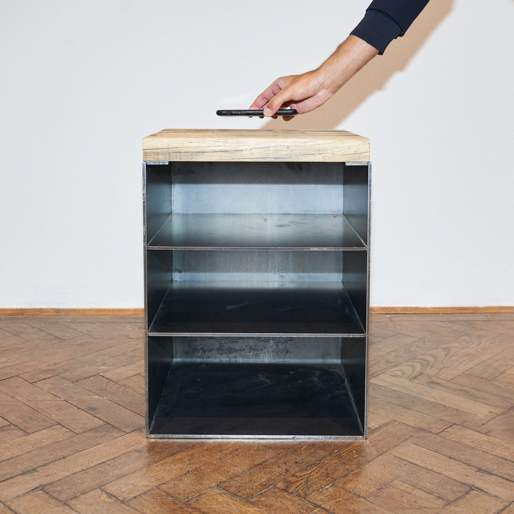 Nachttisch aus rohem Stahl, drei Fächern und einer Platte aus Altholz Fichte, auf die eine Person ein Handy legt